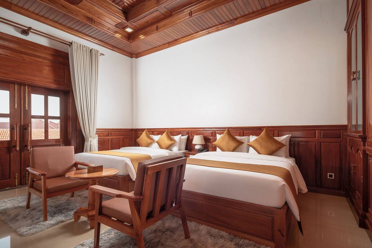 A hotel room with two beds, wooden furniture, a window with curtains, and a wooden ceiling.