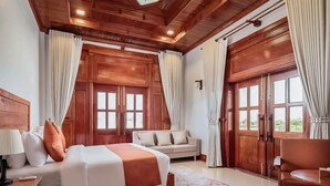 Une chambre spacieuse avec un grand lit, un canapé, une chaise en bois et une vue dégagée sur l’extérieur grâce à une grande fenêtre.