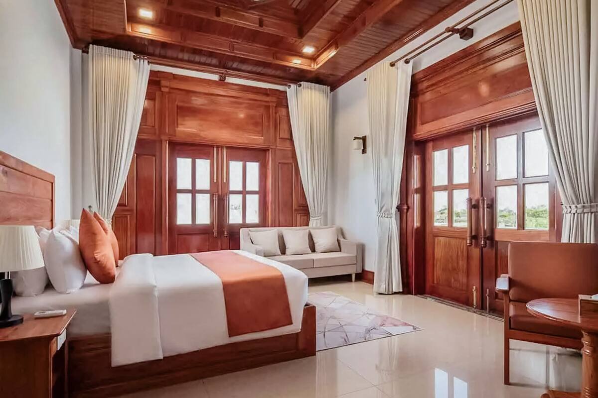 A spacious bedroom with a large bed, a sofa, a wooden chair, and a view through a large window.