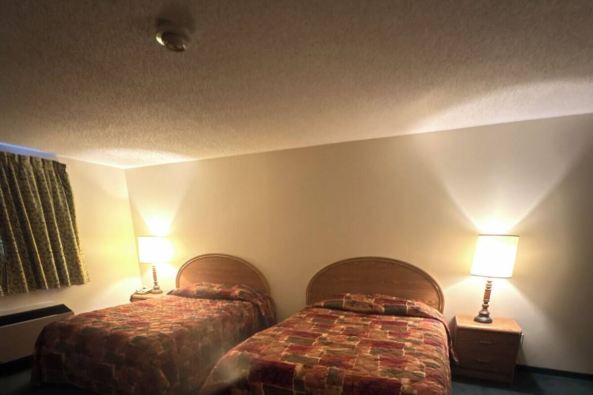 A room with two beds, each with a headboard, a nightstand with a lamp, and a wall-mounted heater.