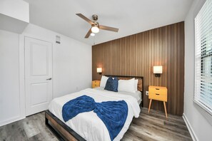 A bedroom with a bed, wooden panel wall, ceiling fan, bedside lamps, and a window with blinds. - Arbor Flats #6 in the Heart of Houston (Houston)