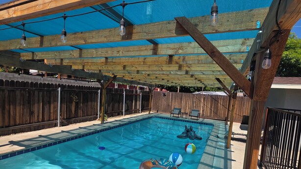 A swimming pool with inflatable rings, a wooden pergola, and a clear blue sky. - Wi-fi / Pool / Ac / Parking / Patio / Grill /Workspace / Ice Chest. (Bakersfield)