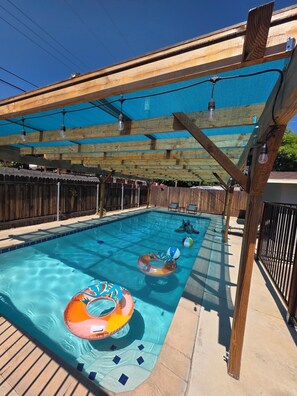 Une piscine avec des bouées gonflables, une pergola en bois et un ciel d’un bleu limpide.