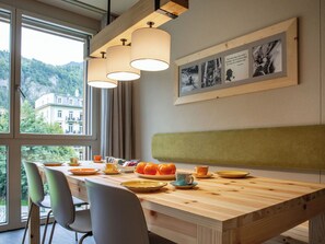 A dining area with a wooden table, chairs, and a bench. There are framed photographs on the wall and a window with a view of buildings and greenery.