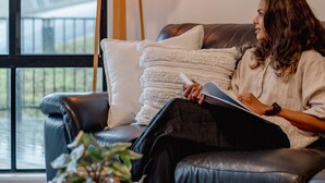 Una persona sentada en un sofá de cuero negro con una manta blanca, leyendo un libro, en una habitación con un gran ventanal, una lámpara de pie y una mesita con una planta.