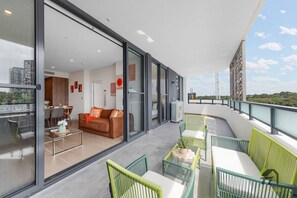 En moderne balkon med glasdøre, der fører til en opholdsstue, udendørs møbler og udsigt over bybilledet.