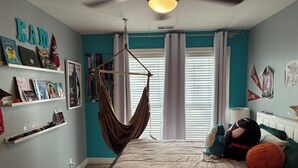 Ein Schlafzimmer mit einem Bett, einer Hängematte, einem Regal mit Büchern und Auszeichnungen sowie einem Fenster mit Vorhängen.