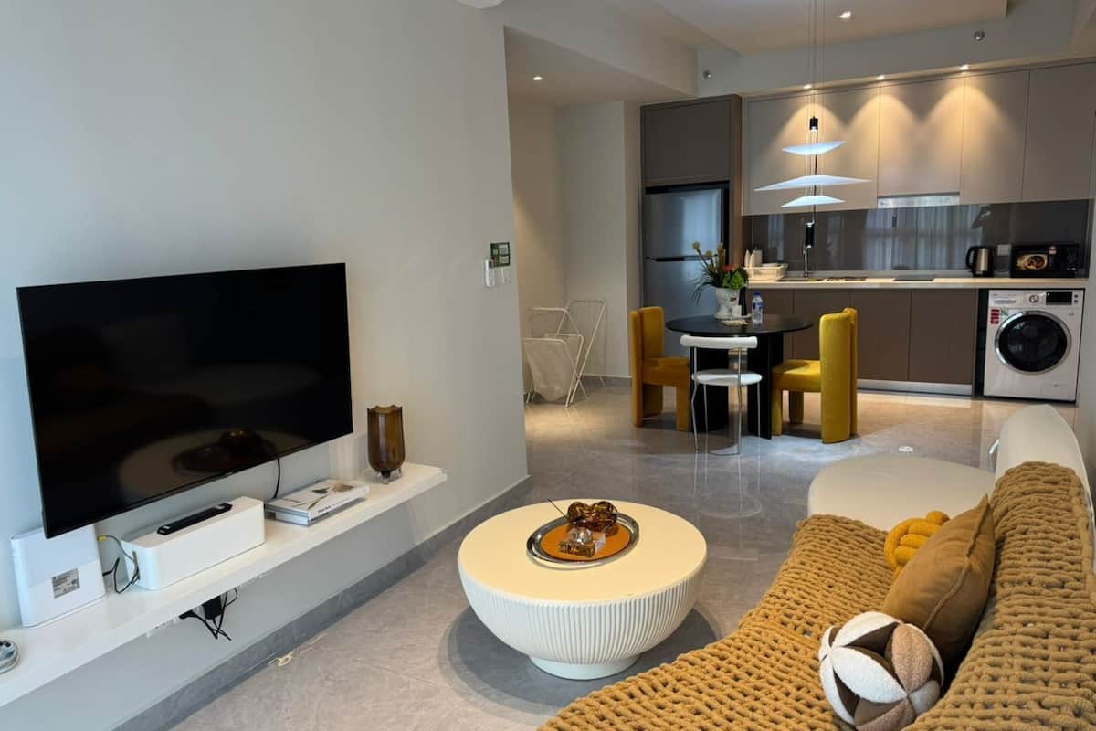 Ein modernes Wohnzimmer mit Flachbildfernseher, rundem Couchtisch, einer Couch und einer Küchenzeile mit Esstisch und Stühlen.