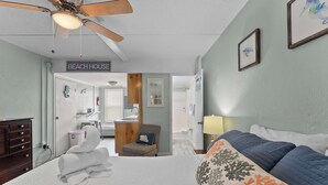 Quarto de hotel com cama, ventilador de teto, placa com a inscrição 'BEACH HOUSE', cadeira com almofada e banheiro visível através de uma porta aberta.