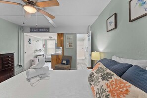 Ein Hotelzimmer mit Bett, Deckenventilator, einem Schild mit der Aufschrift 'BEACH HOUSE', einem Sessel mit Kissen und einem durch eine geöffnete Tür einsehbaren Badezimmer.