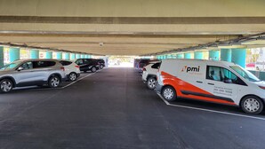 Um parque de estacionamento com veículos, incluindo uma carrinha branca com a marcação 'pmi' e um SUV preto.