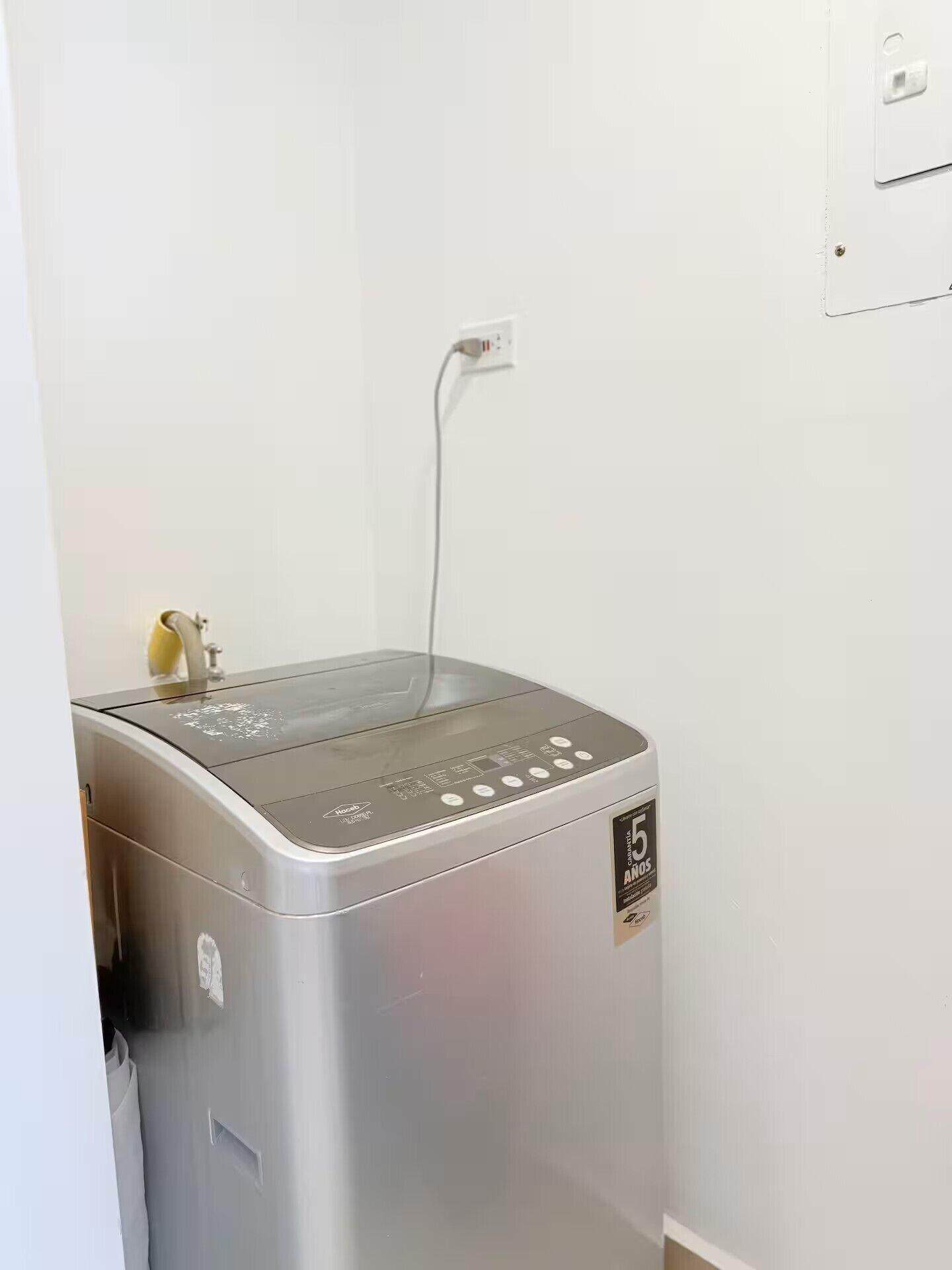 Eine kompakte, silberfarbene, an der Wand befestigte Waschmaschine mit Bedienfeld, einem Stromkabel und einem kleinen, gelben Gegenstand obenauf.