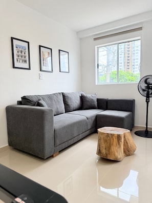 Ein modernes Wohnzimmer mit einem grauen Ecksofa, einem Holztisch, einem Standventilator und gerahmten Bildern an der Wand.