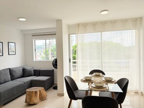 Ein modernes Wohnzimmer mit grauem Sofa, einem runden Holztisch und einem Essbereich mit Glastisch und schwarzen Stühlen.