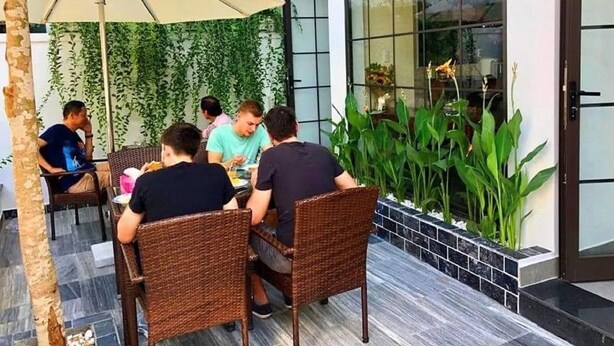 En gruppe mennesker spiser ved et udend ørs bord med fletstole, omgivet af grønne planter og en glasvæg.