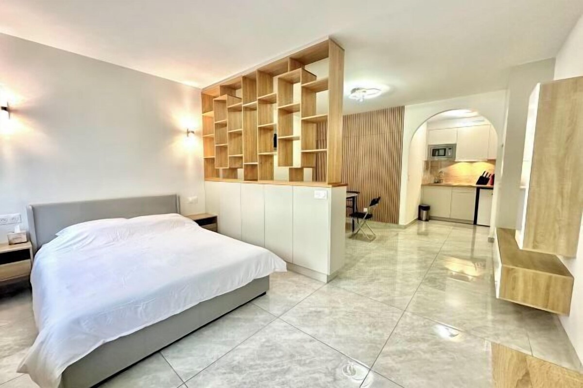 Een moderne hotelkamer met een bed, inbouwplanken, een kitchenette en een badkamer die door een boog zichtbaar is.