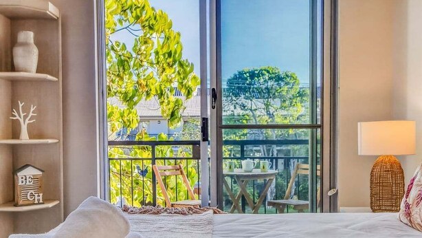 A hotel room with a bed, a balcony with a view of greenery and buildings, a shelf with decorative items, and a lamp. - Coastal Calm at Paradiso - 231 (Tweed)