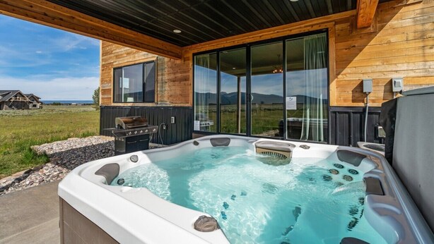 A hot tub with a view of a grassy field and mountains, wooden exterior walls, and a covered patio area.