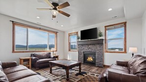 A living room with leather sofas, a fireplace, a ceiling fan, and large windows with a view of the outdoors.