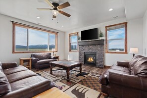 Un salón con sofás de cuero, una chimenea, un ventilador de techo y ventanas grandes con vistas al exterior.