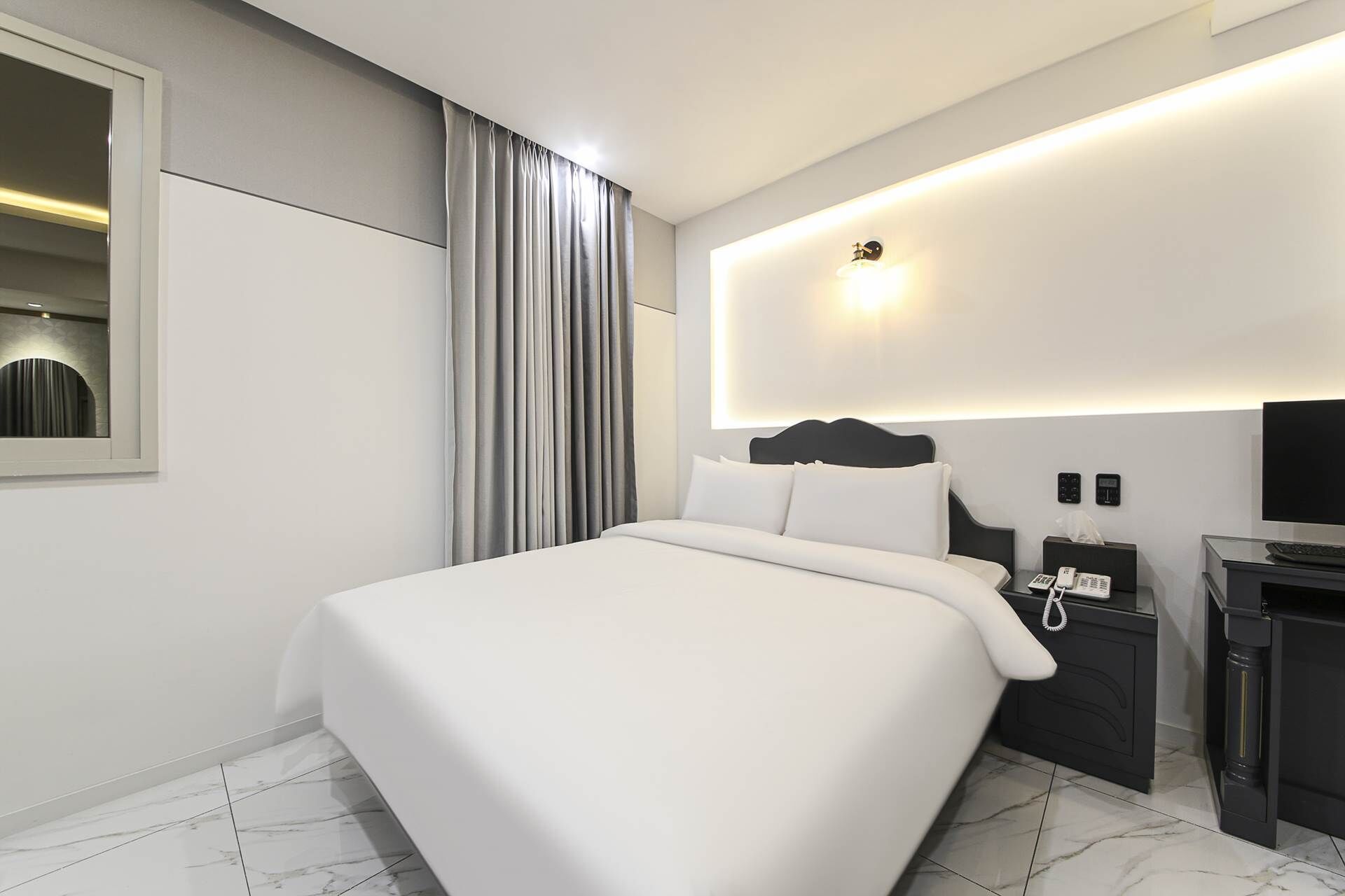 Una habitación de hotel moderna con una cama grande, una mesita de noche con un teléfono y una lámpara, un televisor montado en la pared y un espejo.