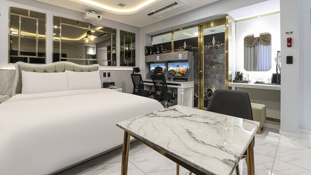 Ein modernes Hotelzimmer mit einem großen Bett, einem Tisch mit Marmorplatte, einem Schreibtisch mit zwei Gaming-Stationen und einer Minibar.
