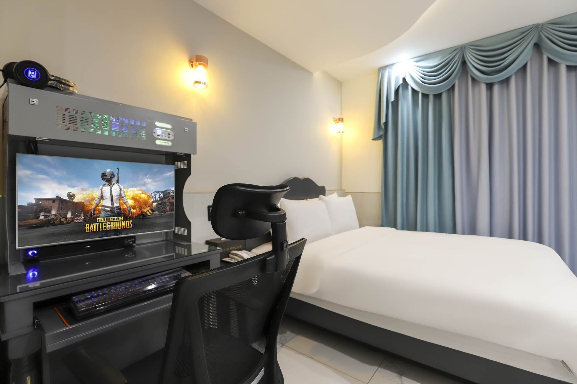 Une chambre d’hôtel avec un lit, un bureau équipé d’un ordinateur, une chaise et un téléviseur diffusant un jeu vidéo.