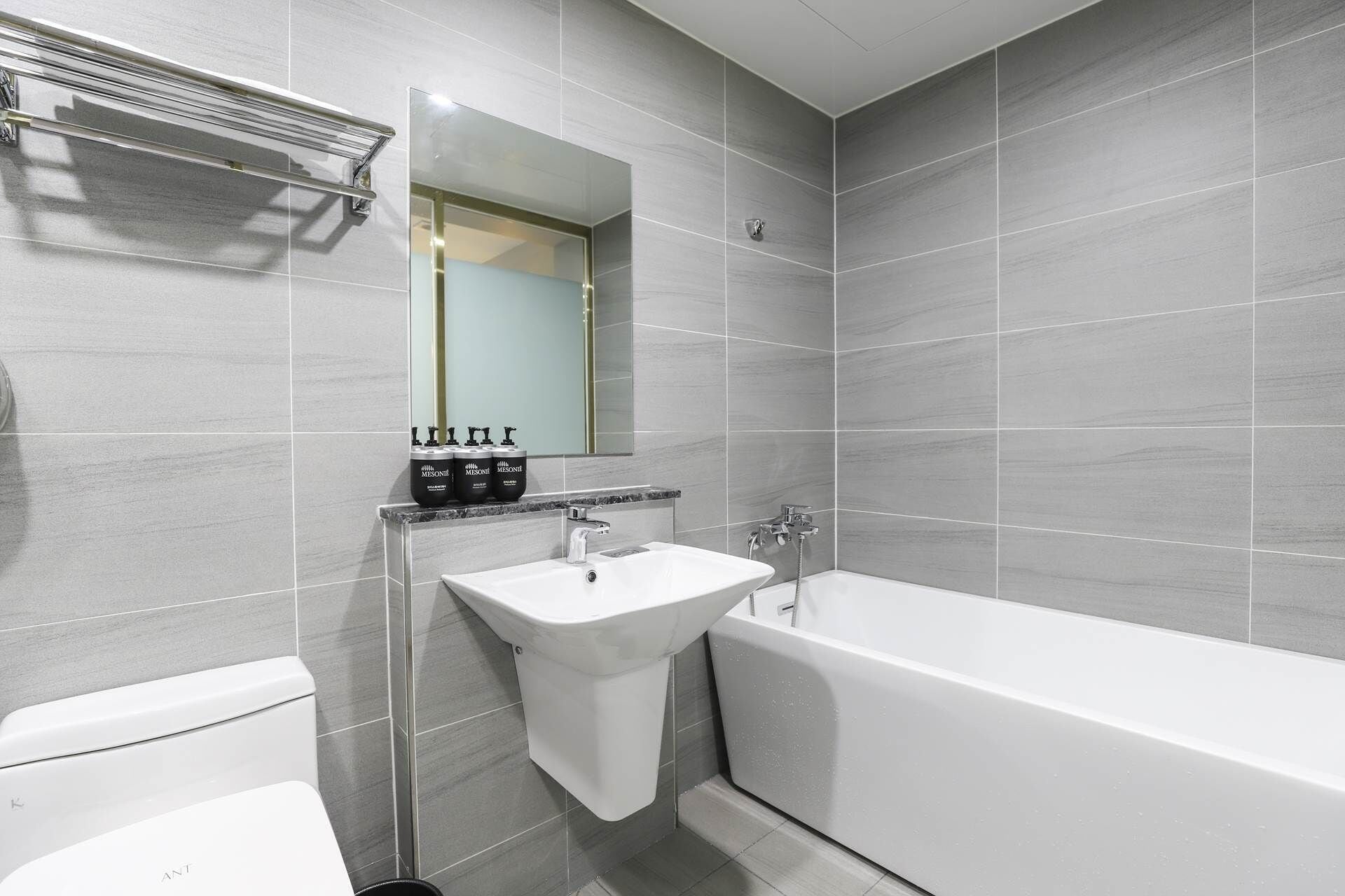 Ein modernes Badezimmer mit einem weißen Waschbecken, einer Badewanne und einer Toilette. Über dem Waschbecken hängt ein Spiegel und darunter befindet sich ein Ablagefach mit Pflegeprodukten.
