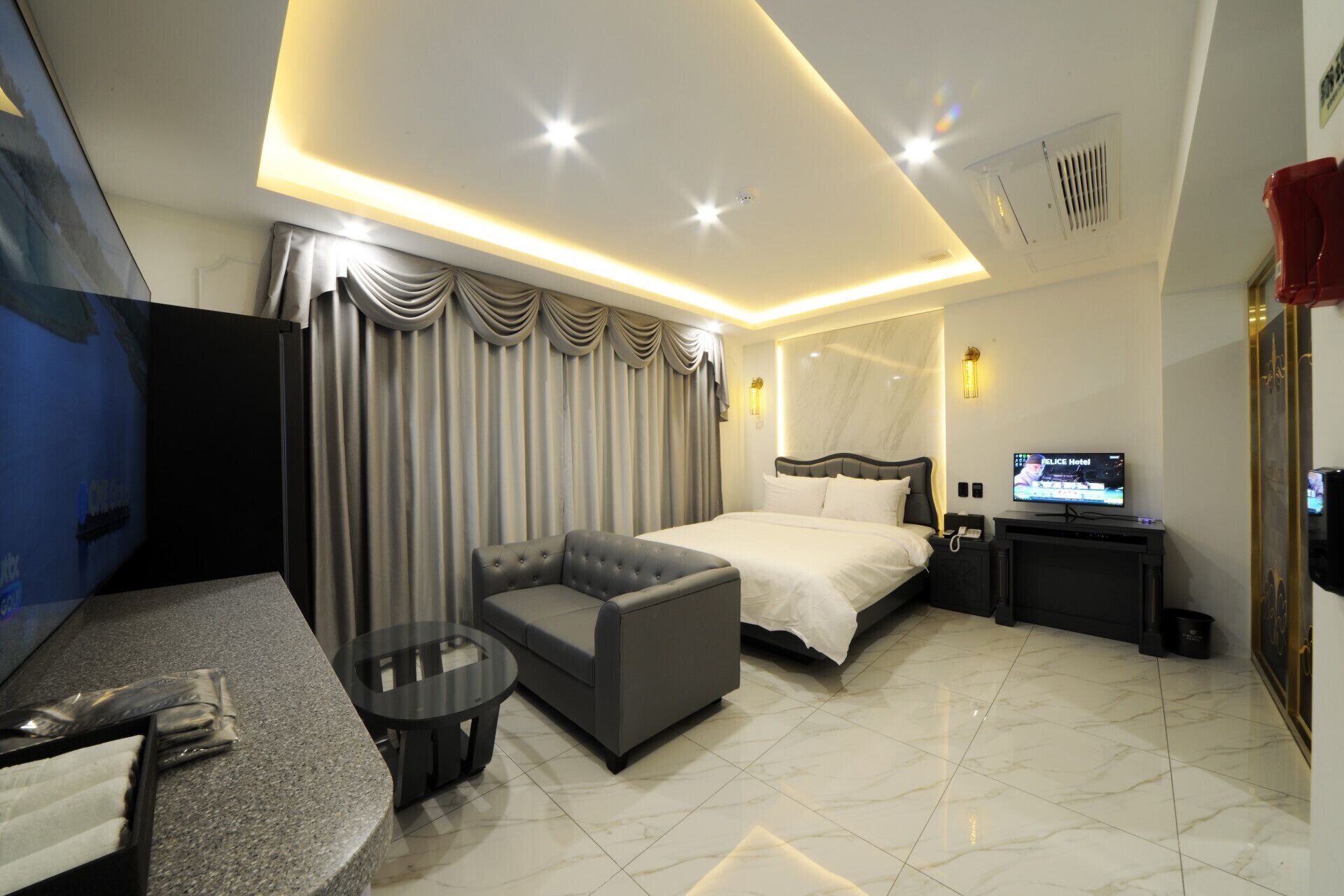 A modern hotel room with a bed, a sofa, a small table, a TV, and a desk with a computer.