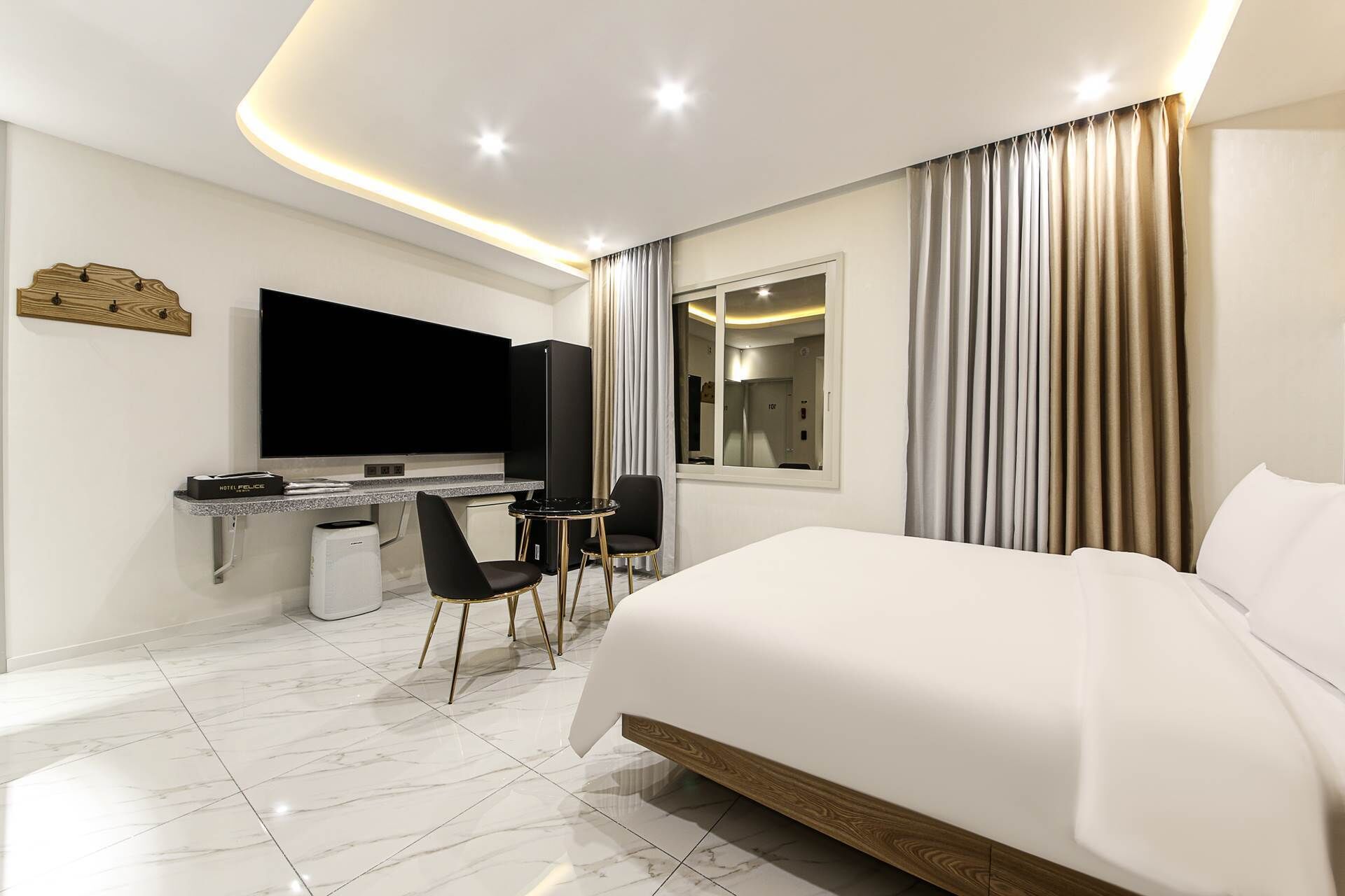 A modern hotel room with a large bed, a desk with a flat-screen TV, a small seating area, and a large window with curtains.