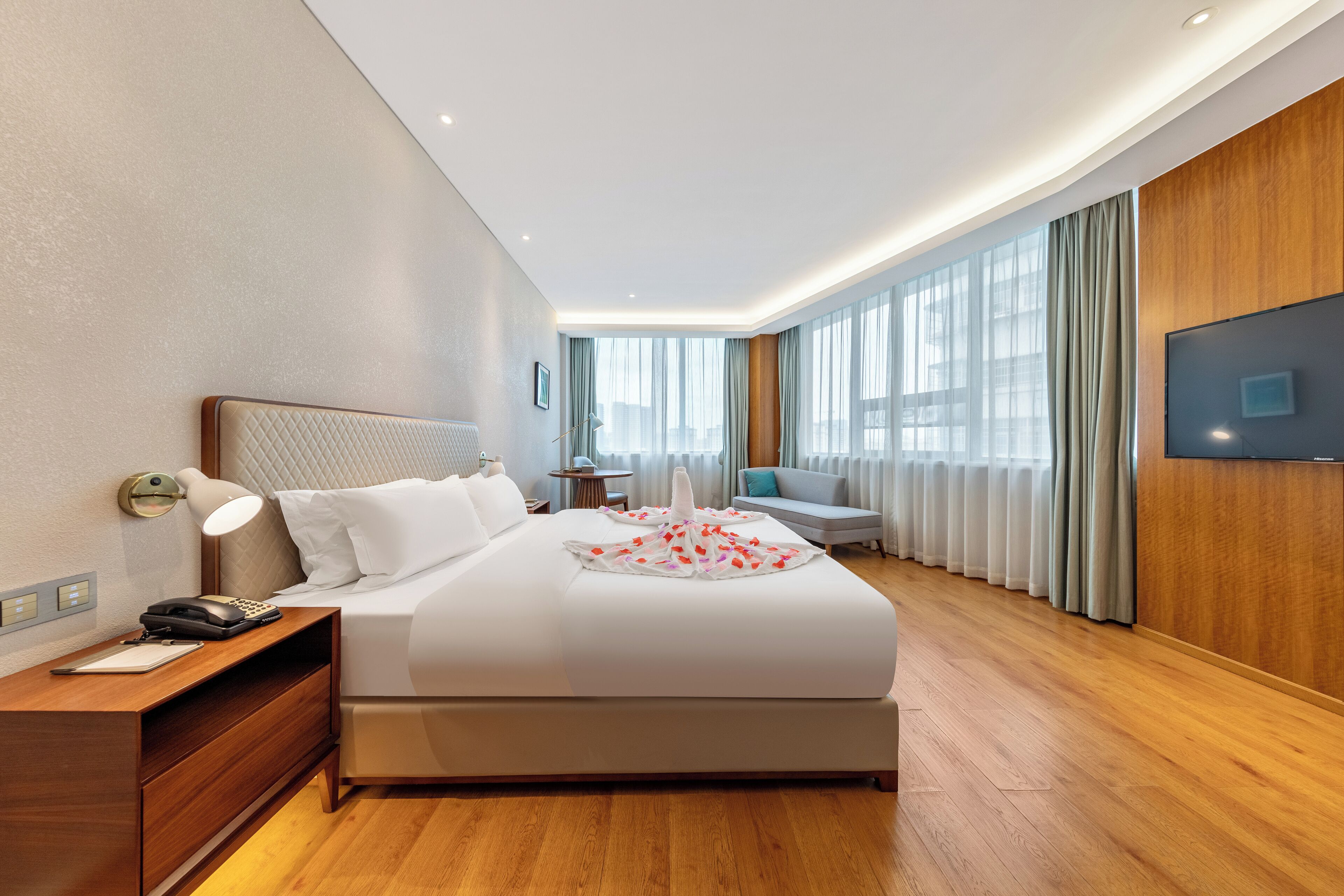 A modern hotel room with a large bed, bedside table, lamp, and a flat-screen TV mounted on the wall.