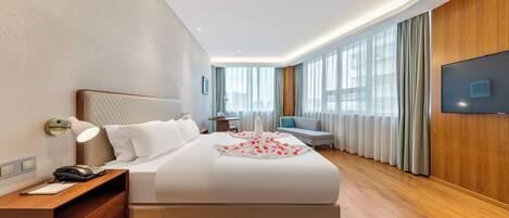 A modern hotel room with a large bed, bedside table, lamp, and a flat-screen TV mounted on the wall.
