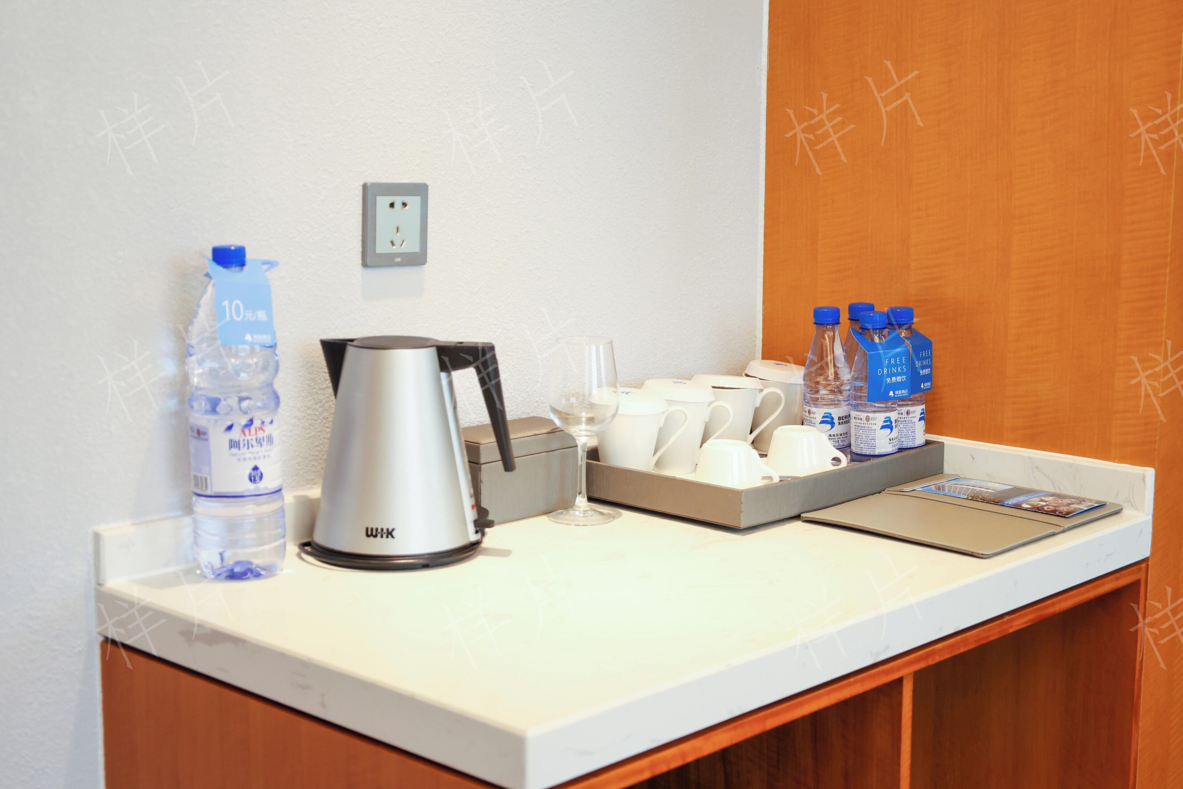 A hotel room counter with a kettle, water bottle, glass, cups, and a tray with bottles.