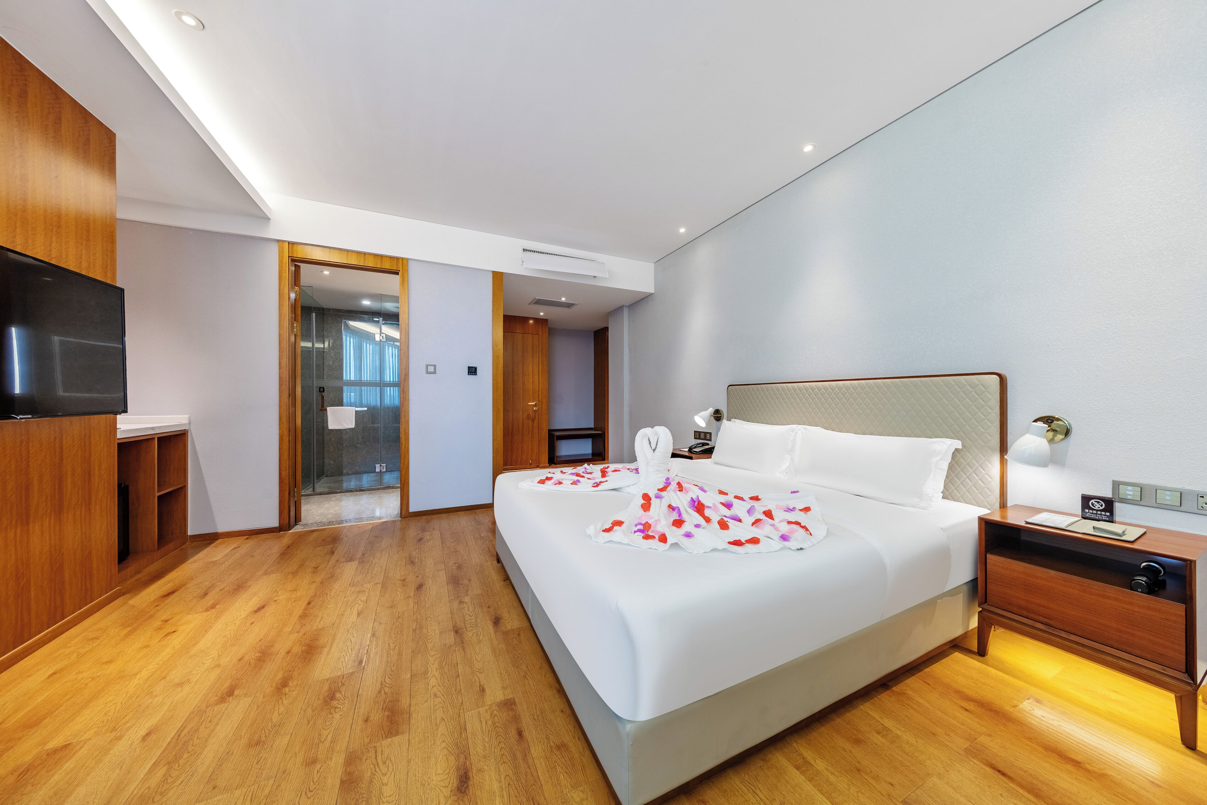 A modern hotel room with a large bed, bedside tables, a TV, and a glass shower area.