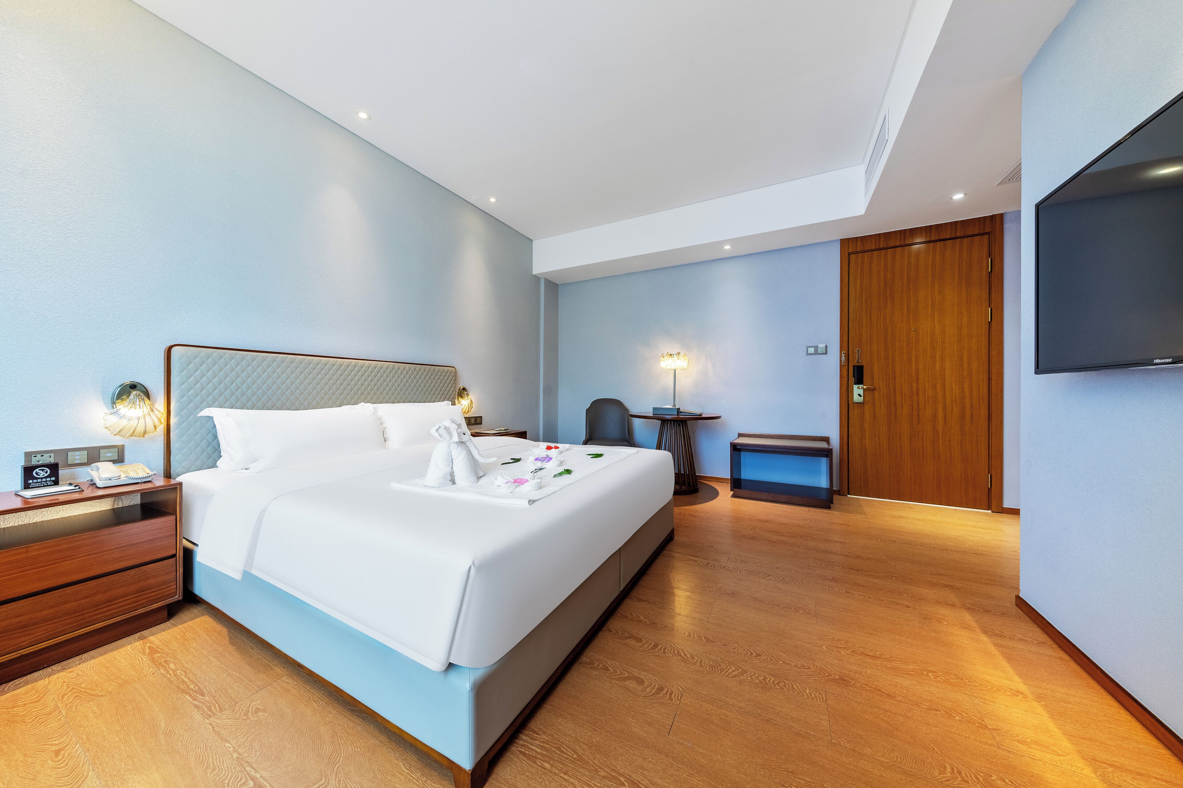 A modern hotel room with a large bed, bedside tables, a desk, a chair, a TV, and a wardrobe.