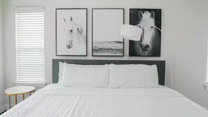 Quarto com cama, roupa de cama branca, mesa de cabeceira redonda e três quadros de cavalos na parede.