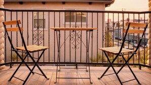 Uma varanda com duas cadeiras de madeira e uma mesa, corrimão e vista para a paisagem urbana.