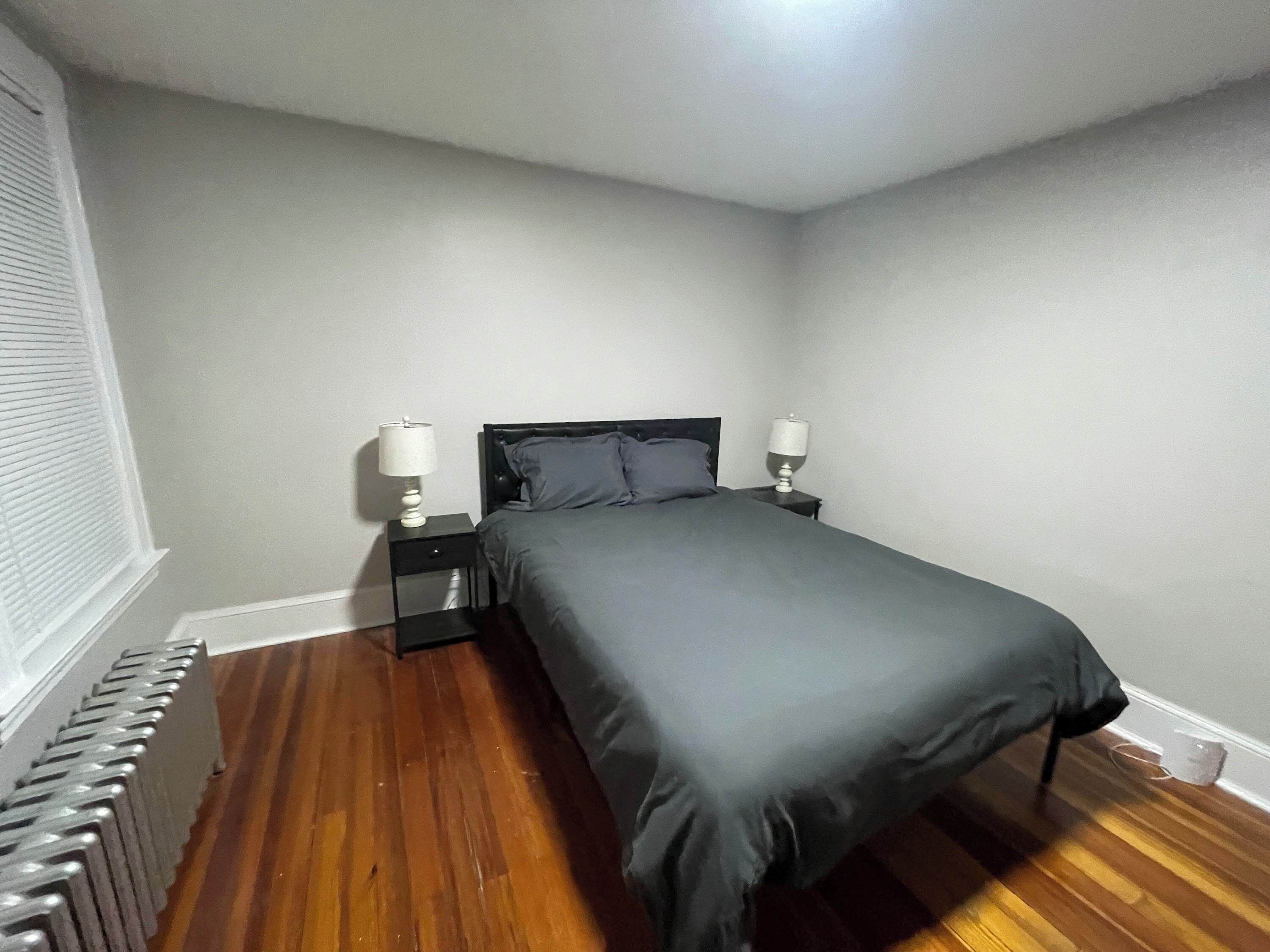 Une chambre à coucher avec un lit, deux tables de chevet avec des lampes, un radiateur et une fenêtre avec des stores.