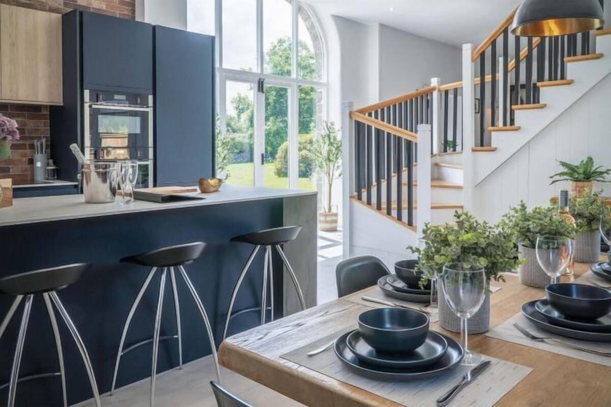 Eine moderne Küche mit Einbauofen, ein Essbereich mit Holztisch und Barhocker sowie eine Treppe, die zu einer oberen Ebene führt.