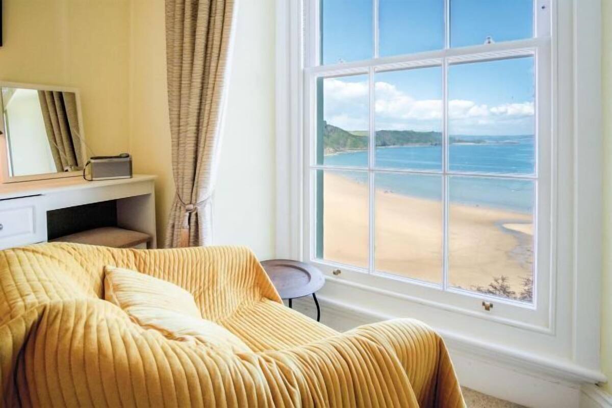 A room with a bed, a window with a beach view, a small table, and a chair.
