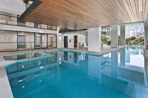 A modern indoor swimming pool with clear water, a wooden ceiling, and a view of the cityscape. - Stylish 1BR Executive Apartment with Pool & Gym (South Brisbane)