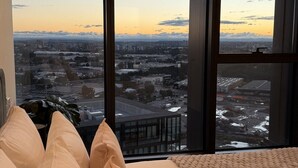 A bedroom with a large window, a bed with beige bedding, and a view of a cityscape at sunset.