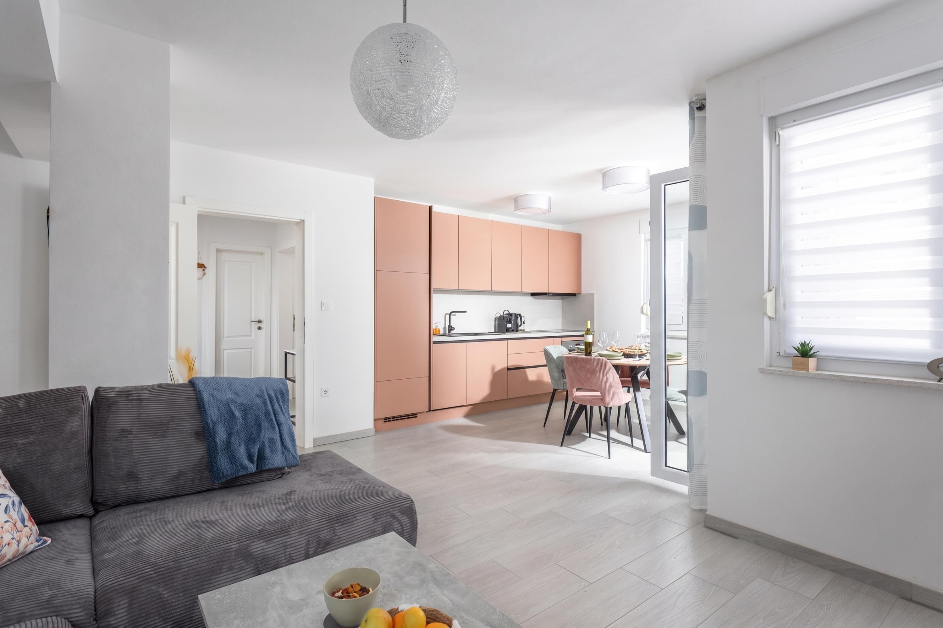 A modern apartment with a grey sofa, a dining area with pink chairs, and a kitchen with peach cabinets.