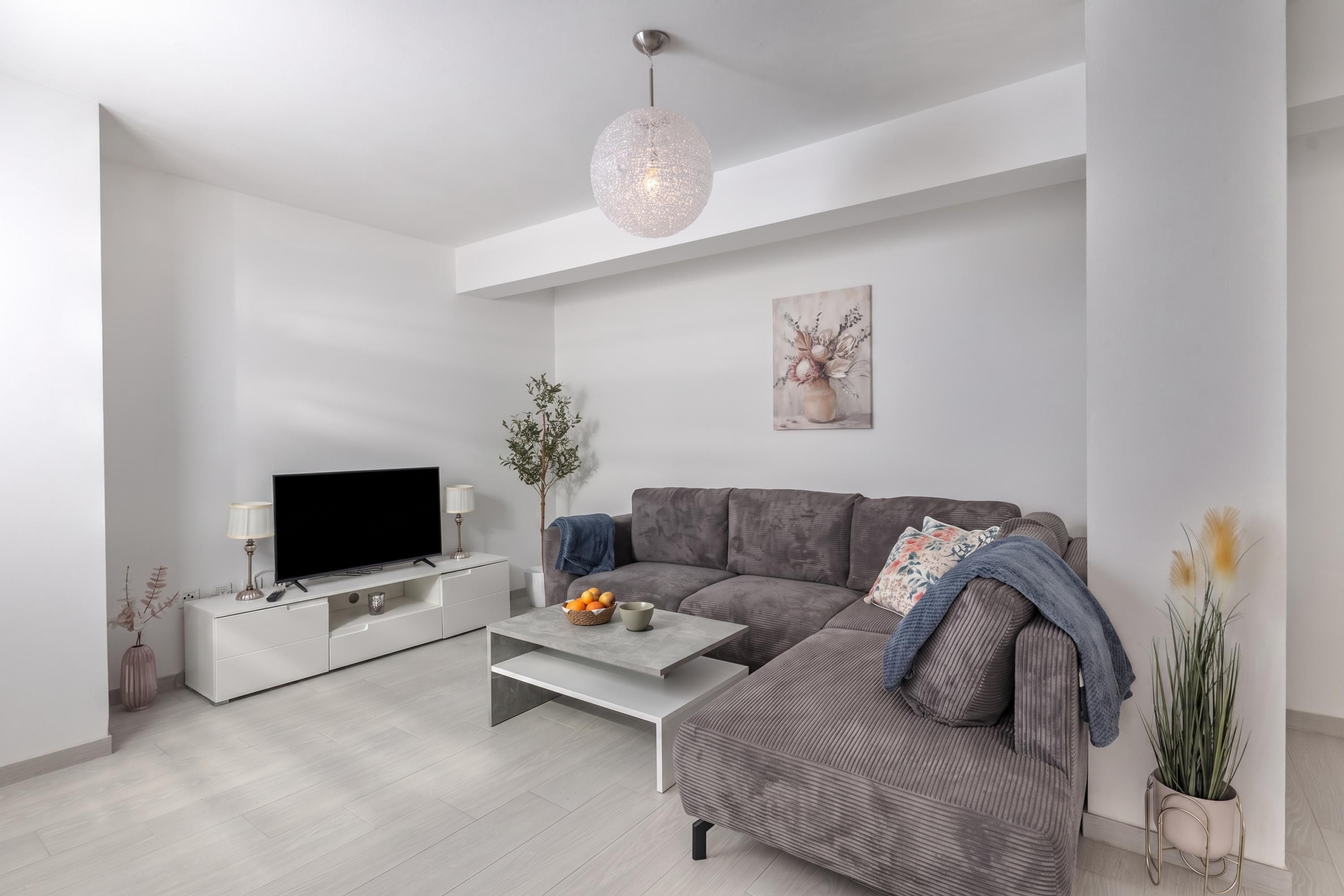 Ein modernes Wohnzimmer mit grauem Sofa, einem Flachbildfernseher auf einem weißen Schrank, einem Couchtisch und einer Pendelleuchte.