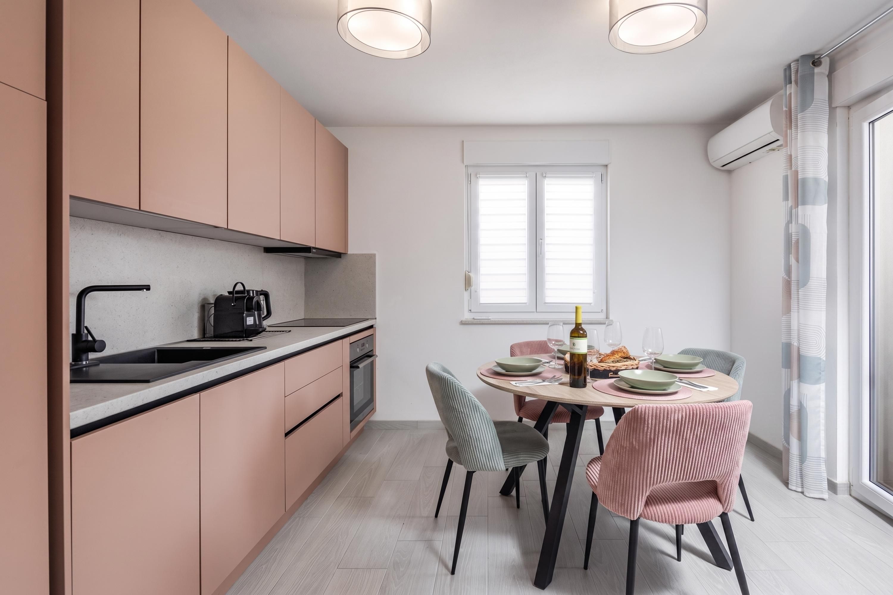 A modern kitchen with a dining area, featuring a round table with chairs, a coffee machine, and a window with blinds.