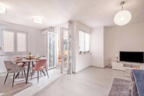 A modern living room with a dining area, a flat-screen TV, and a sliding glass door leading to an outdoor space. - Apt Rosa Walk To Beach /w Balcony (Medulin)