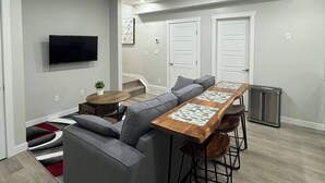 Ein modernes Wohnzimmer mit einer Eckcouch, einem Holzesstisch mit Barhocker, einem flachbildfernseher an der Wand und einer Treppe, die in eine obere Ebene führt.