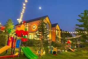 Un bâtiment avec une aire de jeux devant, entouré de verdure et illuminé de guirlandes lumineuses.