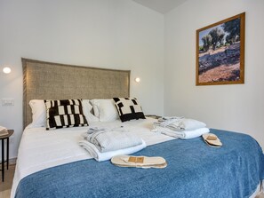 Een keurig opgemaakt bed met kussens, een opgevouwen badjas en pantoffels, een klein tafeltje met een lamp, en een lijst met een foto aan de muur.