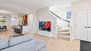 A modern living room with a grey sofa, a TV on a white stand, a kitchen in the background, and a staircase leading to another floor. - Perfect 3-bedroom house in lovely Calgary for your getaway (Calgary)